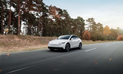 White Tesla Model Y electric SUV driving on scenic highway with forest and autumn trees