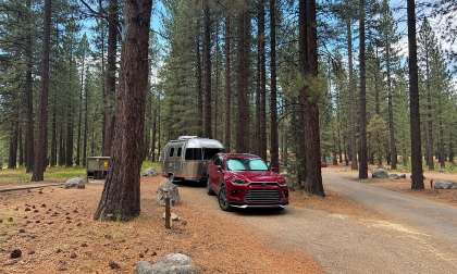 Red Toyota Grand Highlander towing an Airstream travel trailer through a pine forest campsite.