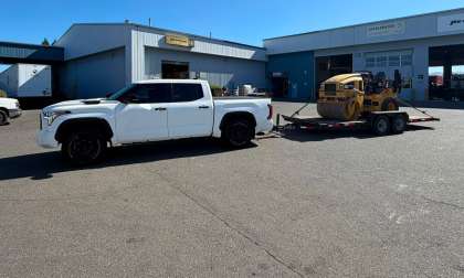 White Toyota Tundra pickup towing a flatbed trailer loaded with a CAT compactor roller outside an industrial equipment yard