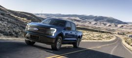 “Blue Ford F-150 Lightning electric truck driving on an open desert highway with mountains in the background.”