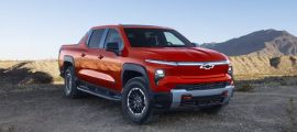 2026 Chevrolet Silverado EV electric pickup truck in red, front three-quarter view off-roading on desert terrain with rugged tires and bold grille design.