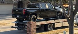 A black Chevrolet Silverado pickup truck being loaded onto a flatbed tow truck, photographed from the side angle in a residential driveway.