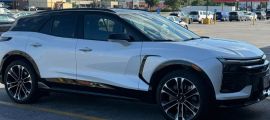 A white Chevrolet Blazer EV is shown from a front three-quarter angle in a parking lot, featuring distinctive black accents and two-tone alloy wheels.