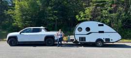 White Chevrolet Silverado EV towing a small white teardrop camper trailer, parked at a forested campsite