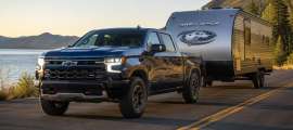 Black Chevrolet Silverado pickup towing a Grey Wolf travel trailer on a road beside a lake and mountains