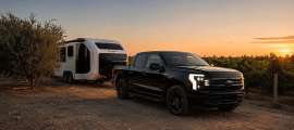 Black Ford F-150 Lightning towing a white Pebble Flow travel trailer through a vineyard at sunset.