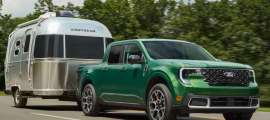 Green Ford Maverick pickup truck towing a silver Airstream travel trailer on a tree-lined highway