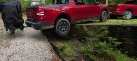 Red Ford Maverick Hybrid XLT towing a covered motorcycle trailer, rear view driving over a low stone retaining wall