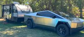 Tesla Cybertruck towing a Patriot Edition travel trailer, parked on grass under shade trees with a dog resting nearby
