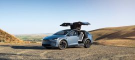 Blue Tesla Model X electric SUV with falcon-wing doors open, parked on a scenic overlook in a rural landscape.