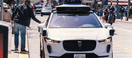 Waymo Jaguar I-Pace robotaxi stopped on a busy city street as a pedestrian passes beside it.