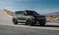 A dark gray/olive 2020 Kia Telluride SUV shown from a front three-quarter angle, driving on a mountain road with black wheels and trim accents against a desert landscape and blue sky.