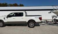 2021 Ford F-150 Platinum in Oxford White, side view, towing a trailer, parked outside white building with metal roof.