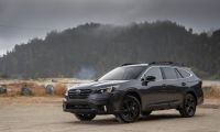 A dark gray 2021 Subaru Outback is shown from a front three-quarter view, parked on a gravel area with misty mountains and forest in the background.