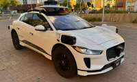 White Jaguar i-Pace with Waymo autonomous driving sensors, parked on brick pavement with urban backdrop