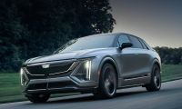 2023 Cadillac Lyriq electric SUV driving on a road, front three-quarter view showing illuminated LED grille and vertical headlights.