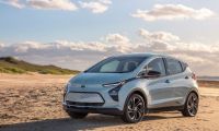 A silver Chevrolet Bolt EV is parked on a sandy beach under a clear blue sky with scattered clouds. The scene conveys a serene and eco-friendly vibe.
