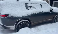 A dark-colored Rivian electric vehicle covered in heavy snow, showing the rear quarter view with illuminated taillights and snow-covered wheels in deep winter conditions.