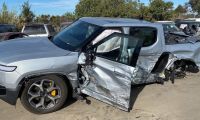 A severely damaged silver Rivian R1T, showing extensive side impact damage and crumpling along its passenger side while parked in what appears to be a salvage yard.