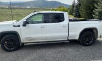 White 2024 GMC Sierra pickup truck with black wheels and off-road tires, photographed from side angle in mountainous rural setting with evergreen trees.