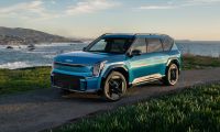 A teal blue 2024 Kia EV9 electric SUV is shown from a front three-quarter angle, parked on a coastal overlook with dramatic ocean waves and mountains in the background.