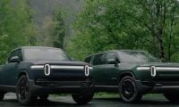 Two 2023 Rivian R1S And Rivian R1T electric pickup trucks in forest green, photographed from front 3/4 view, featuring distinctive LED light bars and black wheels, parked in wooded setting.