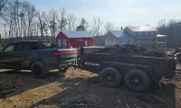 Pickup truck towing a trailer filled with soil, near red and white buildings, surrounded by trees.