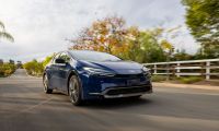 A navy blue 2024 Toyota Prius shown from a front three-quarter angle, driving on a tree-lined suburban road with fall foliage in the background.