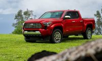 A red 2024 Toyota Tundra pickup truck is shown from a front three-quarter angle, positioned on a grassy hilltop against a cloudy sky backdrop.