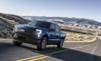 “Blue Ford F-150 Lightning electric truck driving on an open desert highway with mountains in the background.”