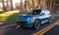 Blue Kia EV9 electric SUV driving on a forest-lined road with motion blur highlighting smooth performance.