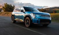Blue 2024 Kia EV9 electric SUV parked on a mountain road with green hills and dramatic clouds in the background.