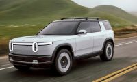 Silver Rivian R2 electric SUV driving on a paved road through rolling green hills with motion blur emphasizing speed.