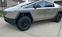 Side view of a Tesla Cybertruck in metallic silver, showcasing its angular design and black off-road wheels with aggressive all-terrain tires parked on a concrete driveway.