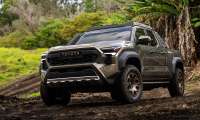 Green 2024 Toyota Tacoma Trailhunter driving off-road on a muddy trail in a front three-quarter view.