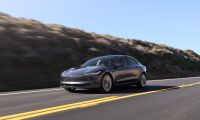 2024 Tesla Model 3 in Midnight Silver Metallic driving on a coastal highway, front three-quarter view with hillside and blue sky background