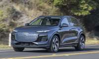 2024 Audi Q6 e-tron in metallic gray, front three-quarter view, driving on winding mountain road with distinctive LED headlights and large octagonal grille.