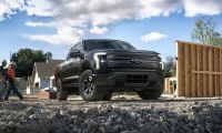 A black Ford F-150 Lightning EV pickup truck, likely a -2022 model, is shown from the front quarter angle at a construction site with wooden fencing panels and rocky ground visible.