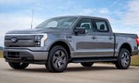 2022 Ford F-150 Lightning electric pickup truck in silver, three-quarter front view, featuring distinctive LED light bar and closed grille, parked on concrete with rural landscape background.