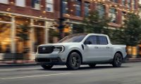 A white 2023 Ford Maverick pickup truck shown from a front three-quarter angle, driving on a city street with motion blur and brick buildings in the background.