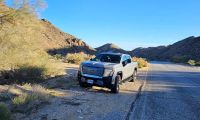 2025 GMC Sierra EV electric pickup truck parked on a desert roadside, showing front three-quarter view with rugged terrain and mountains in the background