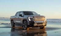 2025 GMC Sierra EV Denali pickup truck in silver parked on a reflective beach at sunset, showcasing its futuristic LED headlights and bold electric truck design.