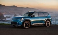 Blue 2025 Kia EV9 electric SUV parked near an ocean cliff at sunset, front three-quarter view.