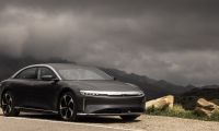 A black Lucid Air electric luxury sedan shown from a front three-quarter angle, parked on a mountain road with dramatic cloudy skies in the background.