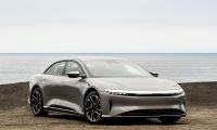 A silver Lucid Air luxury electric sedan shown from a front three-quarter angle, parked on a coastal terrain with the ocean visible in the background.
