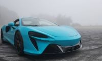 A turquoise 2025 McLaren Artura parked on a misty road, showcasing sleek lines and bold black wheels.