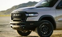A silver 2025 RAM 1500 pickup truck shown from the front quarter angle, featuring black off-road wheels, aggressive honeycomb grille design, and rugged front bumper against a mountainous backdrop.