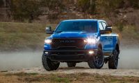 A bright blue 2025 Ram 1500 pickup truck shown from a front three-quarter angle, with LED headlights illuminated and off-road tires kicking up dust on rugged terrain.