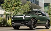 Forest green Rivian R1S electric SUV shown from a front three-quarter angle, parked on a city street with modern architecture and greenery in the background.