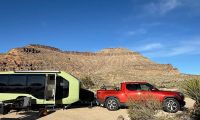 A red Rivian R1T pickup truck is parked next to a lime green camper trailer in a desert landscape with rocky mountains in the background, photographed from a side angle.